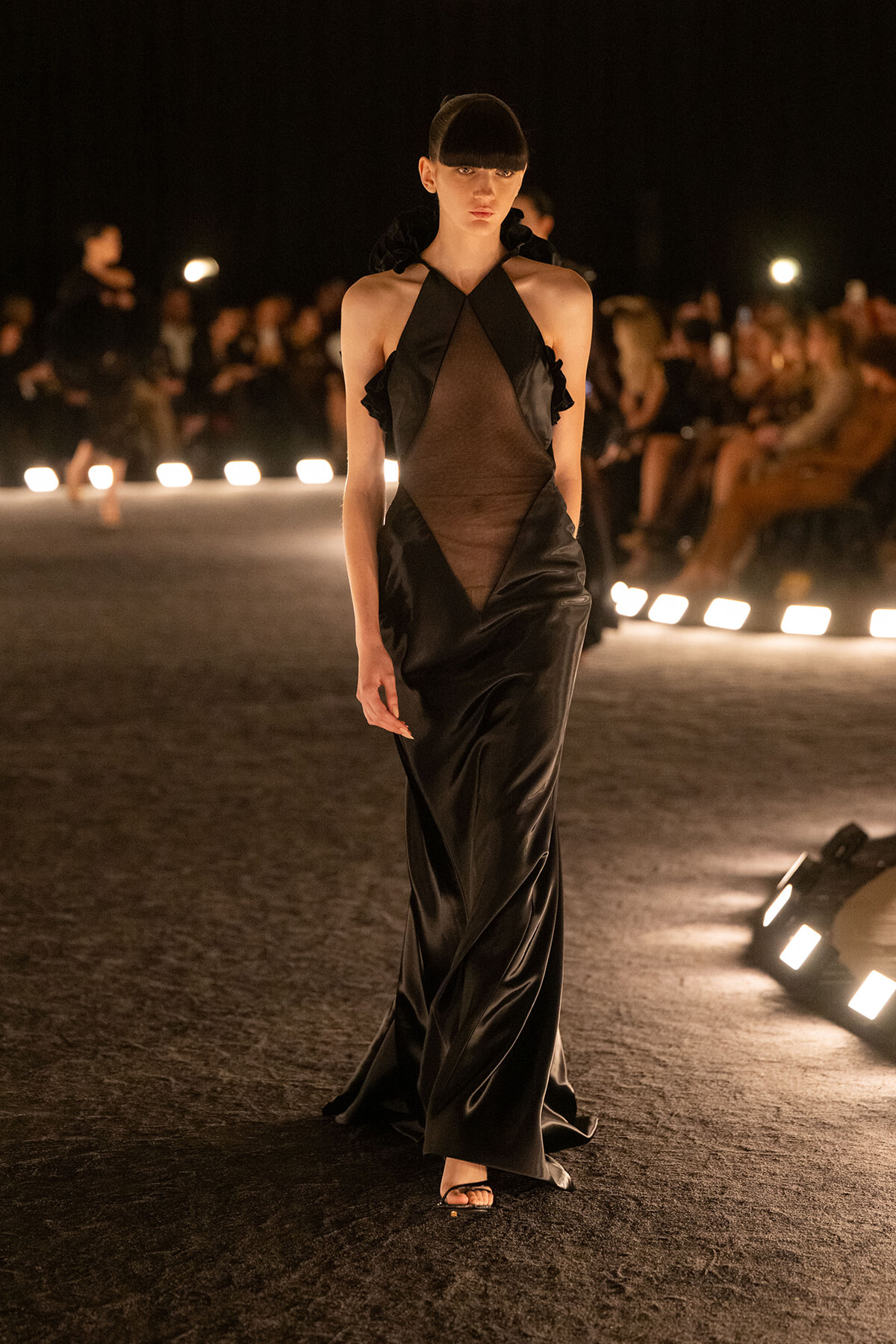 Model wearing a long black satin gown with sheer panels and halter neck walks a nighttime runway; audience blurred in the background.