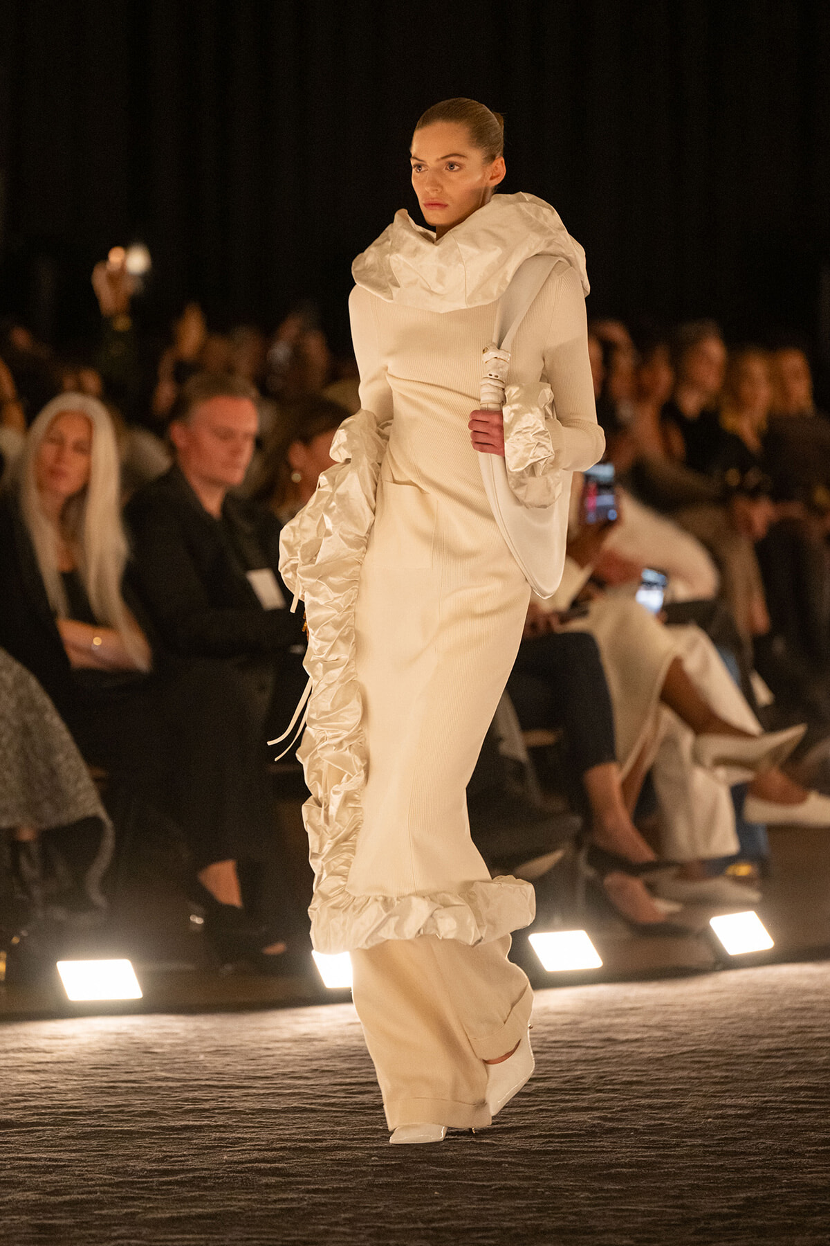 Model on a runway wearing an all-white sculptural outfit with large ruffled collar and sleeves, surrounded by an attentive audience.