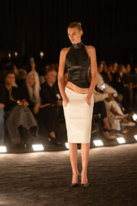 Model walks the runway in a black sleeveless high-neck top with studded cups and a white midi skirt, hands in pockets, under warm runway lighting.