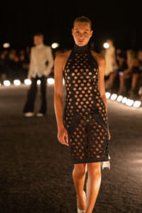 Model walks the runway at night wearing a black perforated halter dress with circular cutouts, spotlighting on the catwalk.
