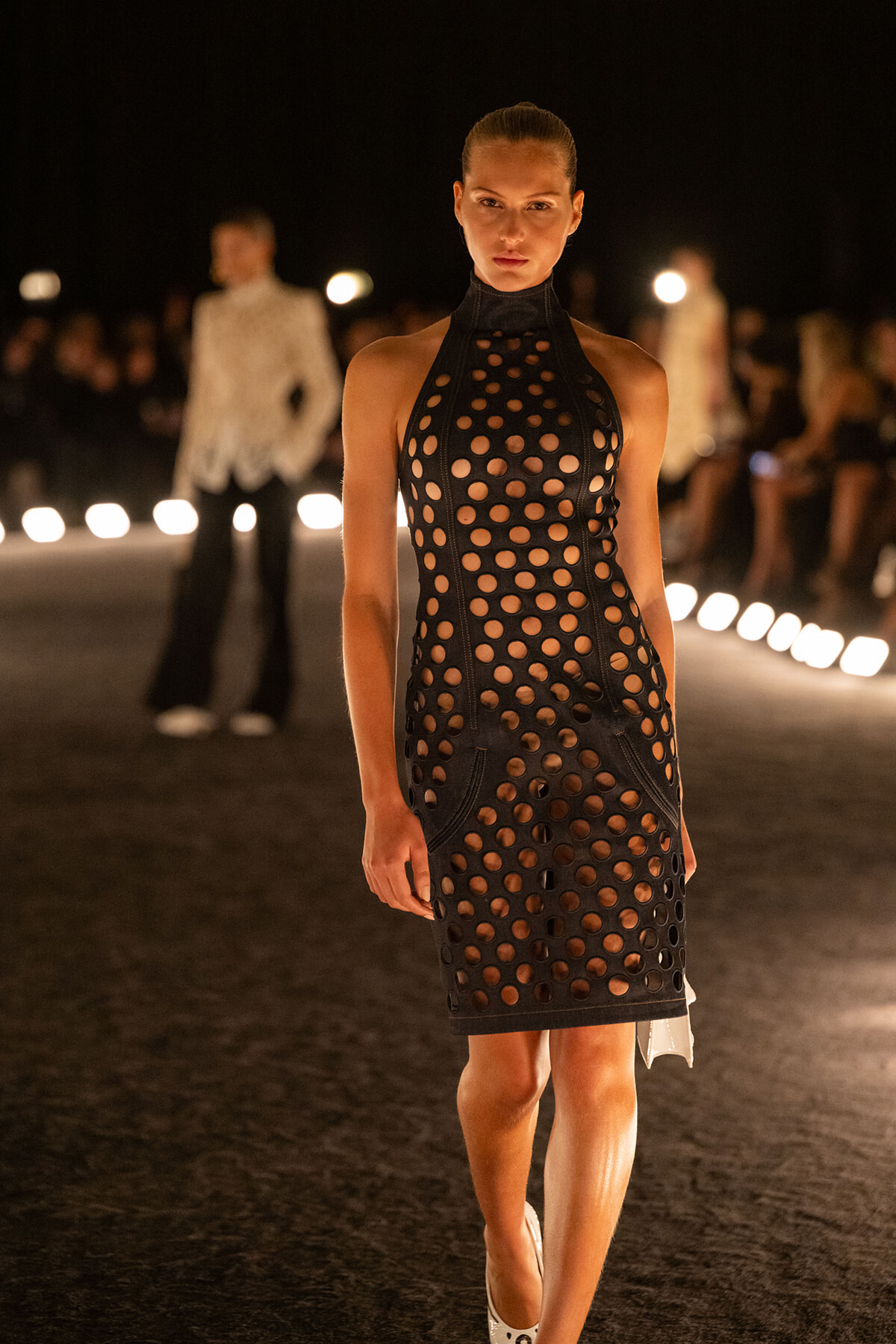 Model walks the runway at night wearing a black perforated halter dress with circular cutouts, spotlighting on the catwalk.