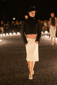 Model walking a fashion runway in a black cropped jacket, white pencil skirt, and a black hat, hands in pockets as audience watches.