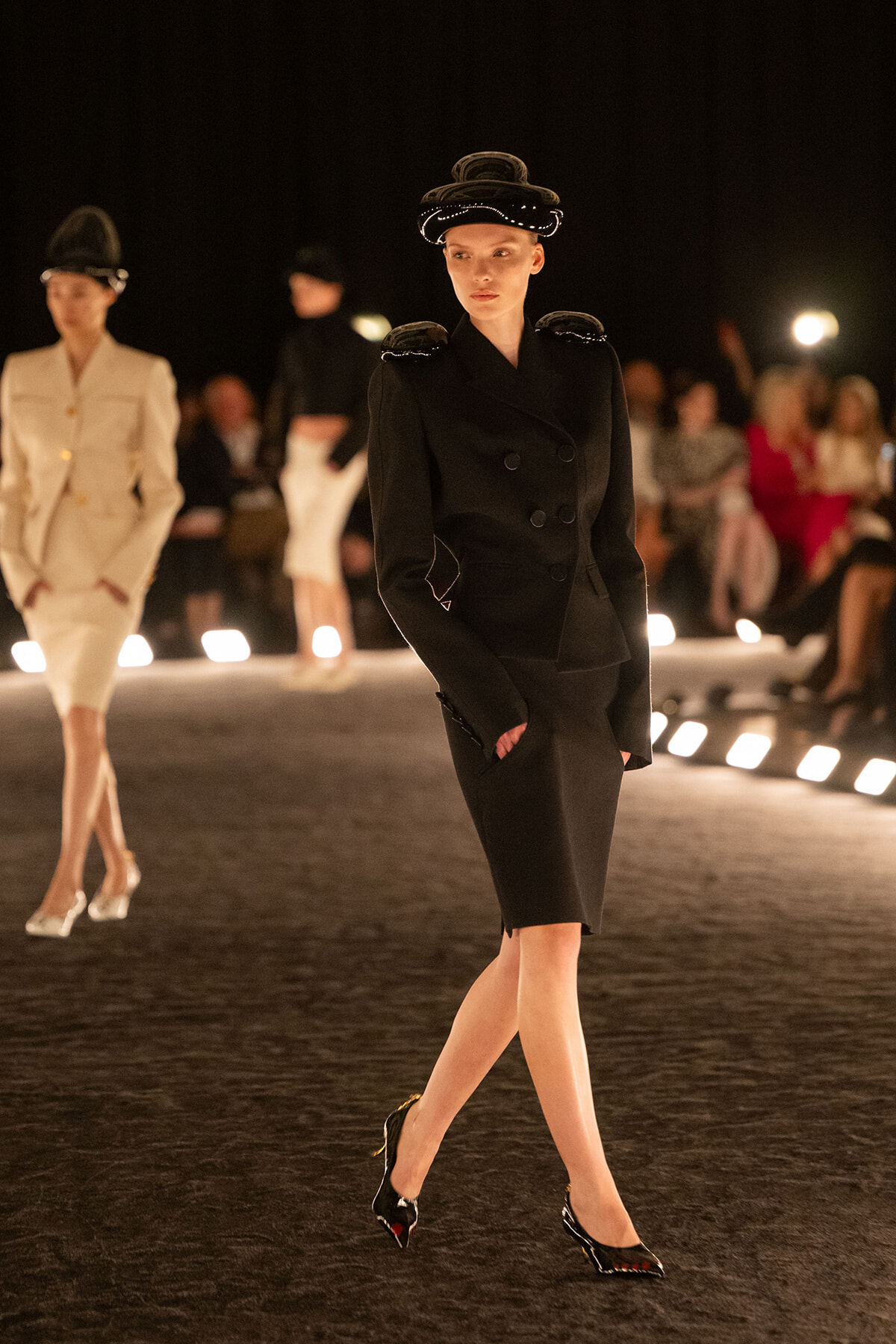 Model on a dim runway wearing a black, double-breasted tailored blazer with matching pencil skirt and ornate hat; patent heels complete the look.