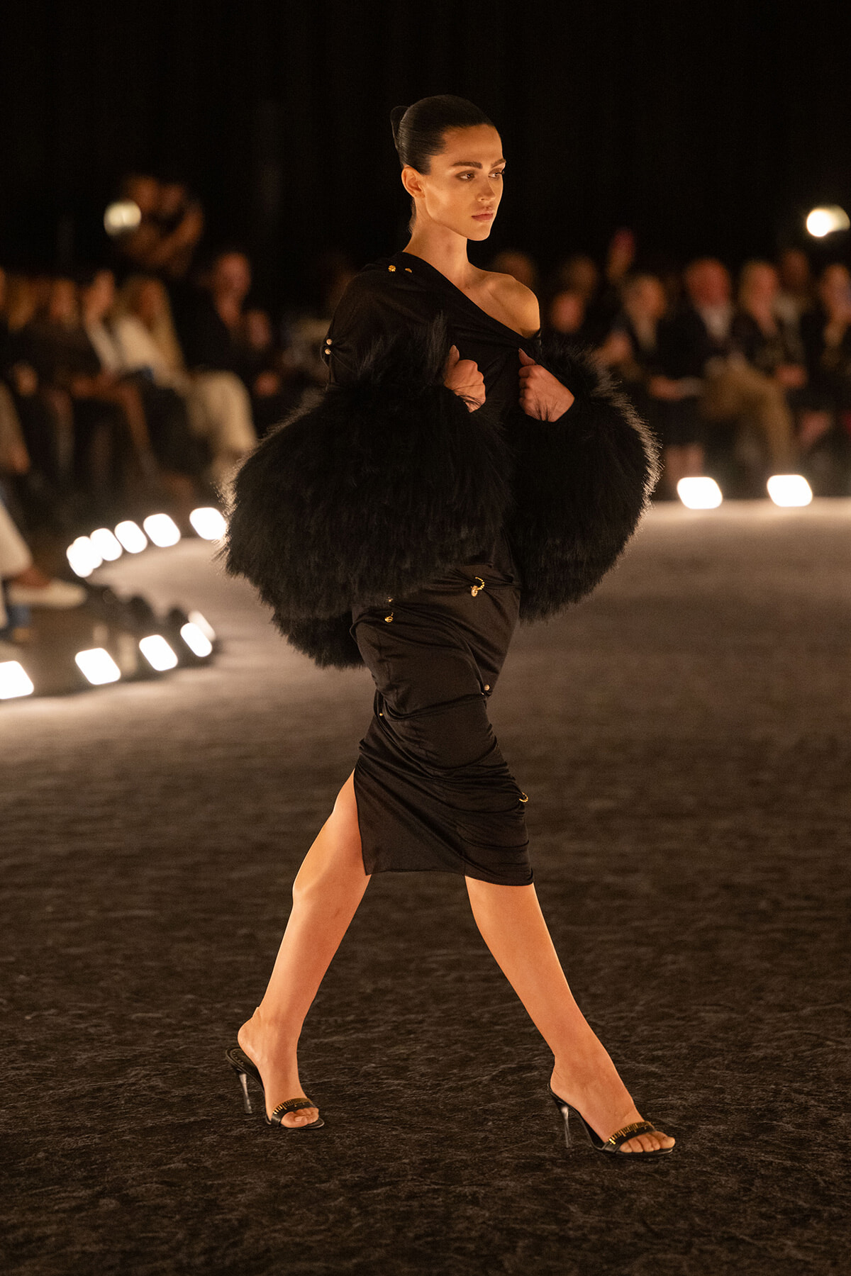 Model walking a runway at night in a black, off-shoulder dress with oversized feathered sleeves and clear heels, audience in the background.