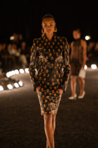 Model walking a runway in a black dotted suit with orange polka dots and matching skirt, bold hoops, spotlighted at night.