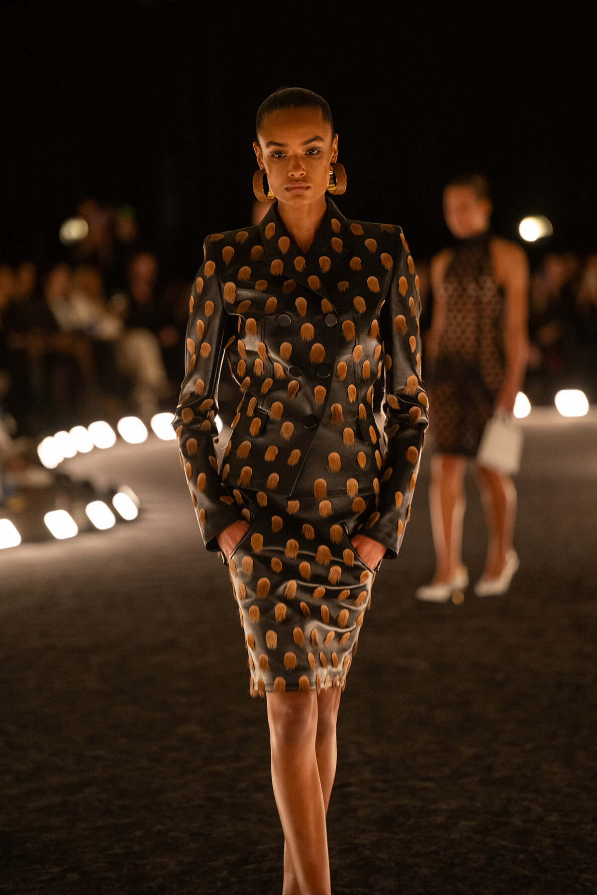Model walking a runway in a black dotted suit with orange polka dots and matching skirt, bold hoops, spotlighted at night.
