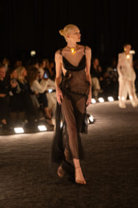 Model walking a dimly lit runway in a sheer black gown with gold necklace, audience in the background.
