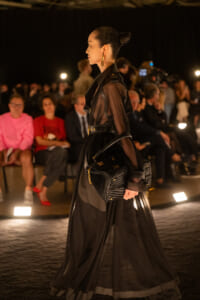 Model walks a runway in a sheer black outfit with a structured belt, gold hoop earrings, and a woven black handbag on a nighttime fashion show stage.