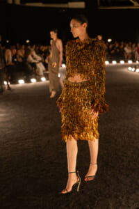 Model wearing a gold fringe two‑piece dress on a runway, walking with an audience in the background at a fashion show.