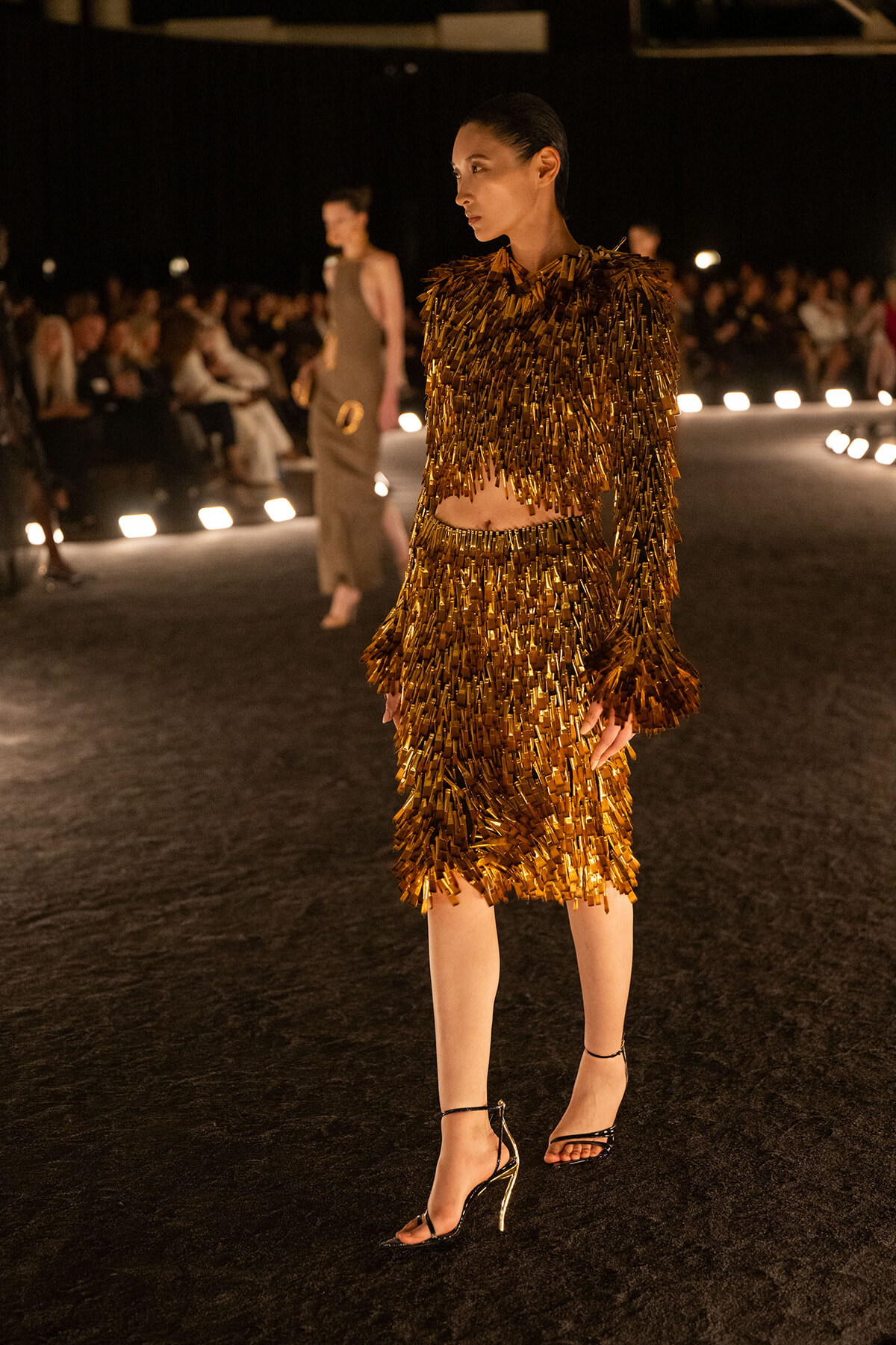 Model wearing a gold fringe two‑piece dress on a runway, walking with an audience in the background at a fashion show.