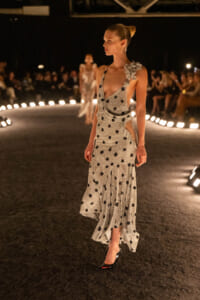 Model walks a backstage-lit runway in a silver sequined polka-dot gown with side slits and a floral shoulder embellishment, black heels on dark carpet.