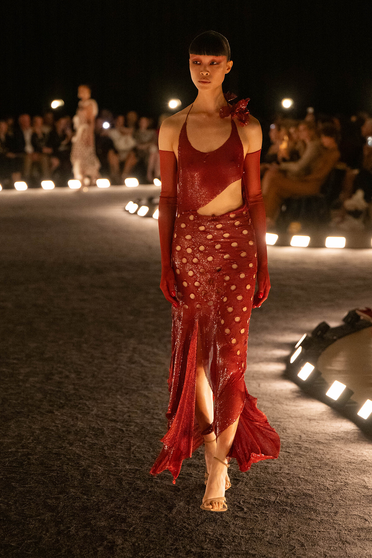 Model walking a red sequined gown with cutouts on a nighttime runway show.