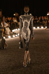 Model on a runway wearing a black dress with bold white abstract pattern, carrying a small handbag.