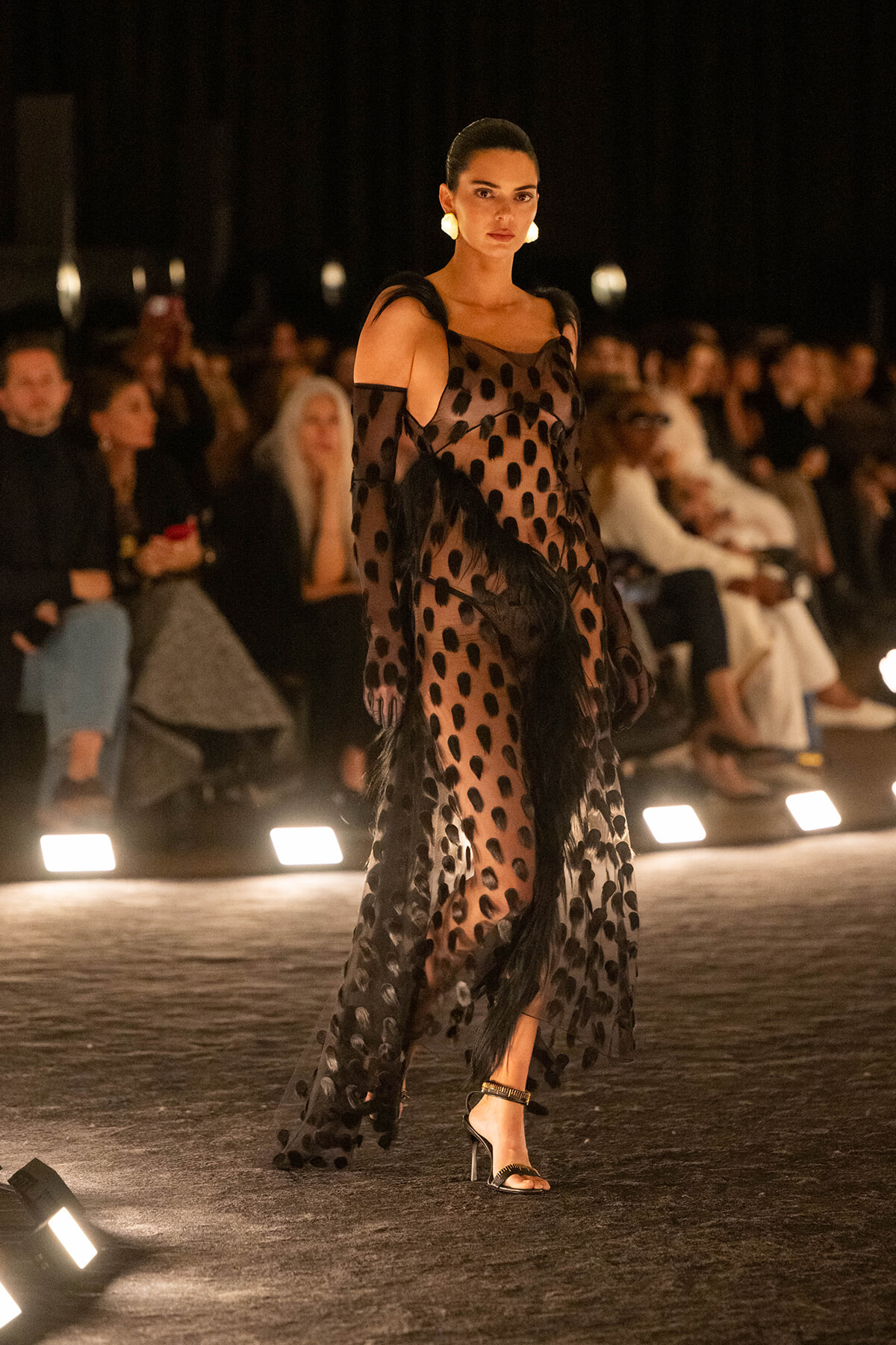 Model wearing a sheer black polka-dot gown with feather details walks the fashion runway.