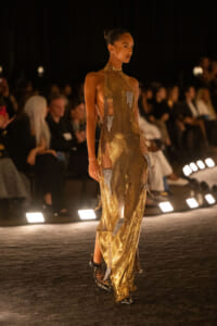 Model wearing a gold sequined halter gown with cutouts, walking a runway under bright spotlights, black ankle boots visible.