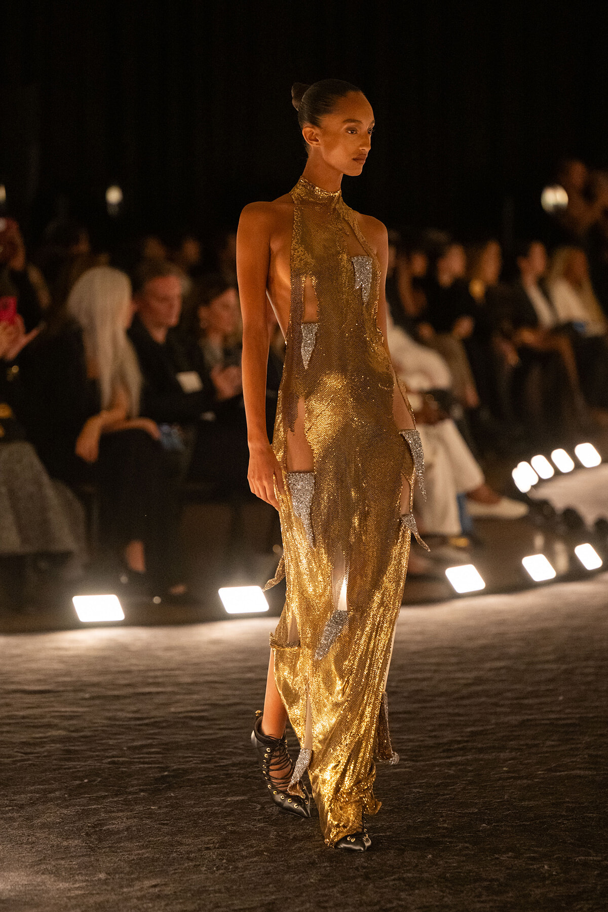 Model wearing a gold sequined halter gown with cutouts, walking a runway under bright spotlights, black ankle boots visible.