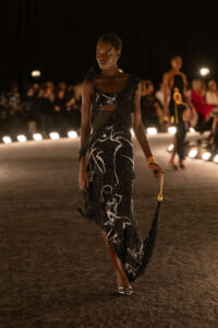 Model walking a nighttime runway in a black sequined gown with white abstract patterns, holding a small gold-chain purse.