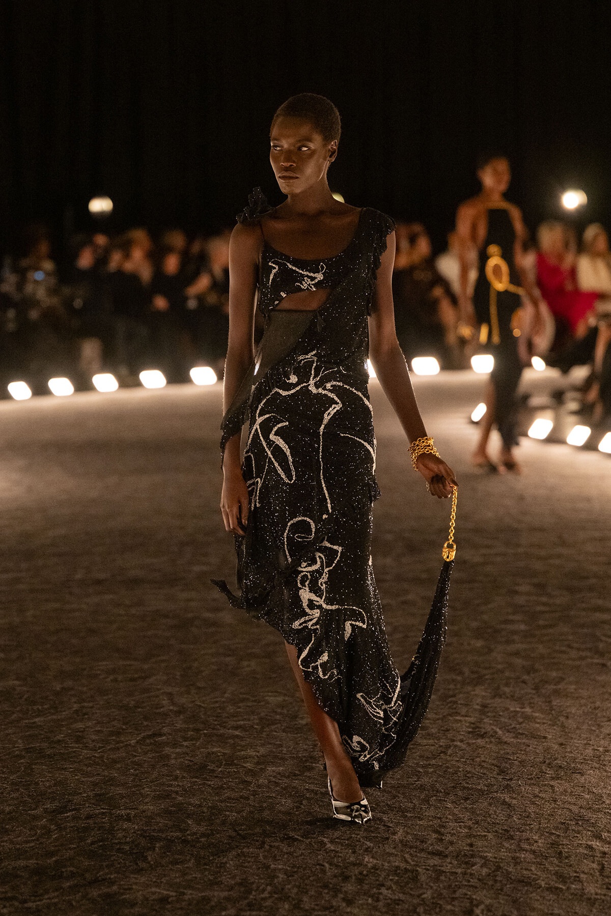 Model walking a nighttime runway in a black sequined gown with white abstract patterns, holding a small gold-chain purse.
