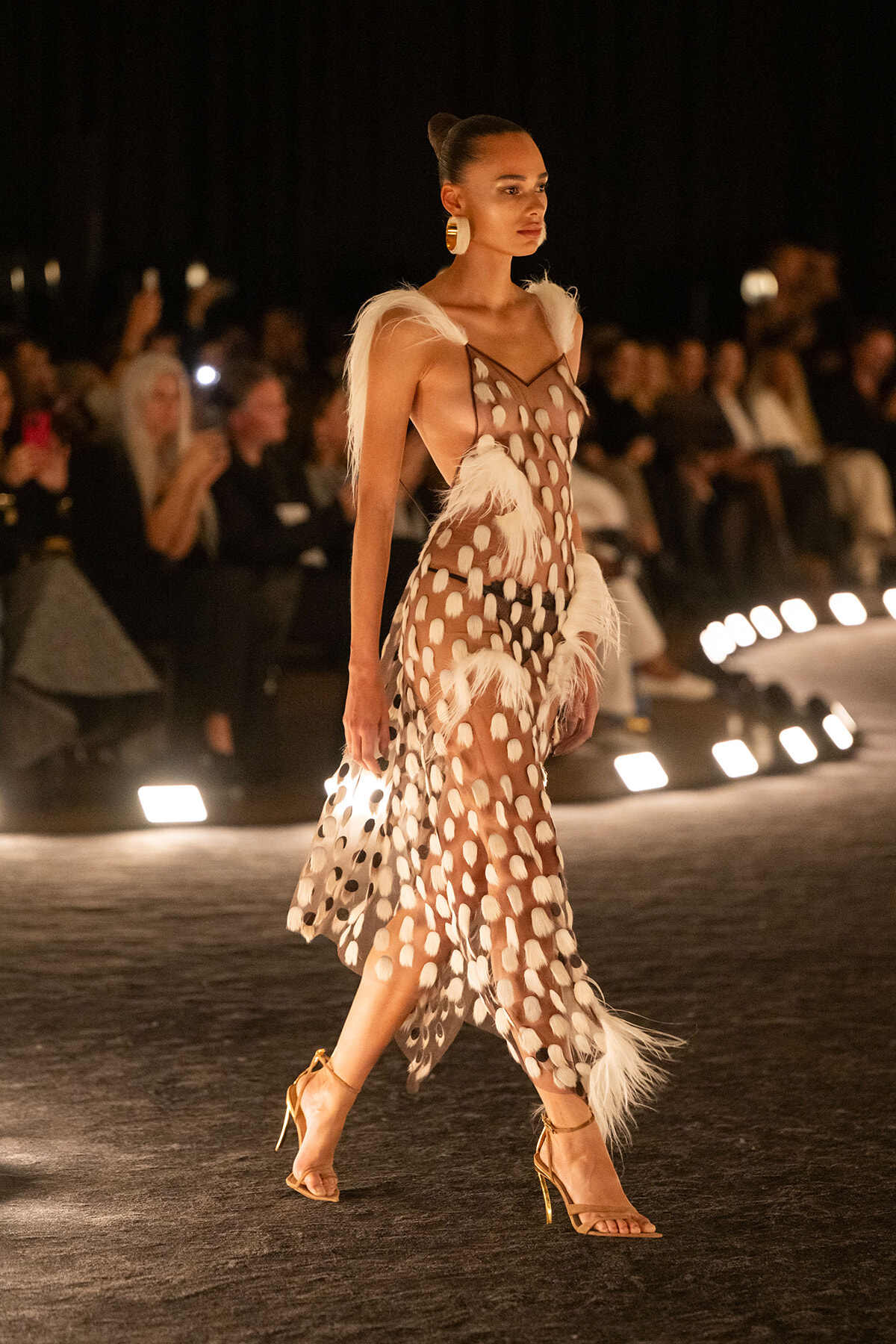 Model walking a nighttime runway in a sheer brown polka-dot dress with white feather trims and gold strappy heels, audience in the background
