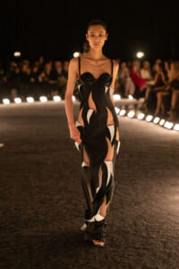Model walking a dimly lit runway in a black cut-out gown with white abstract panels on a dark catwalk.