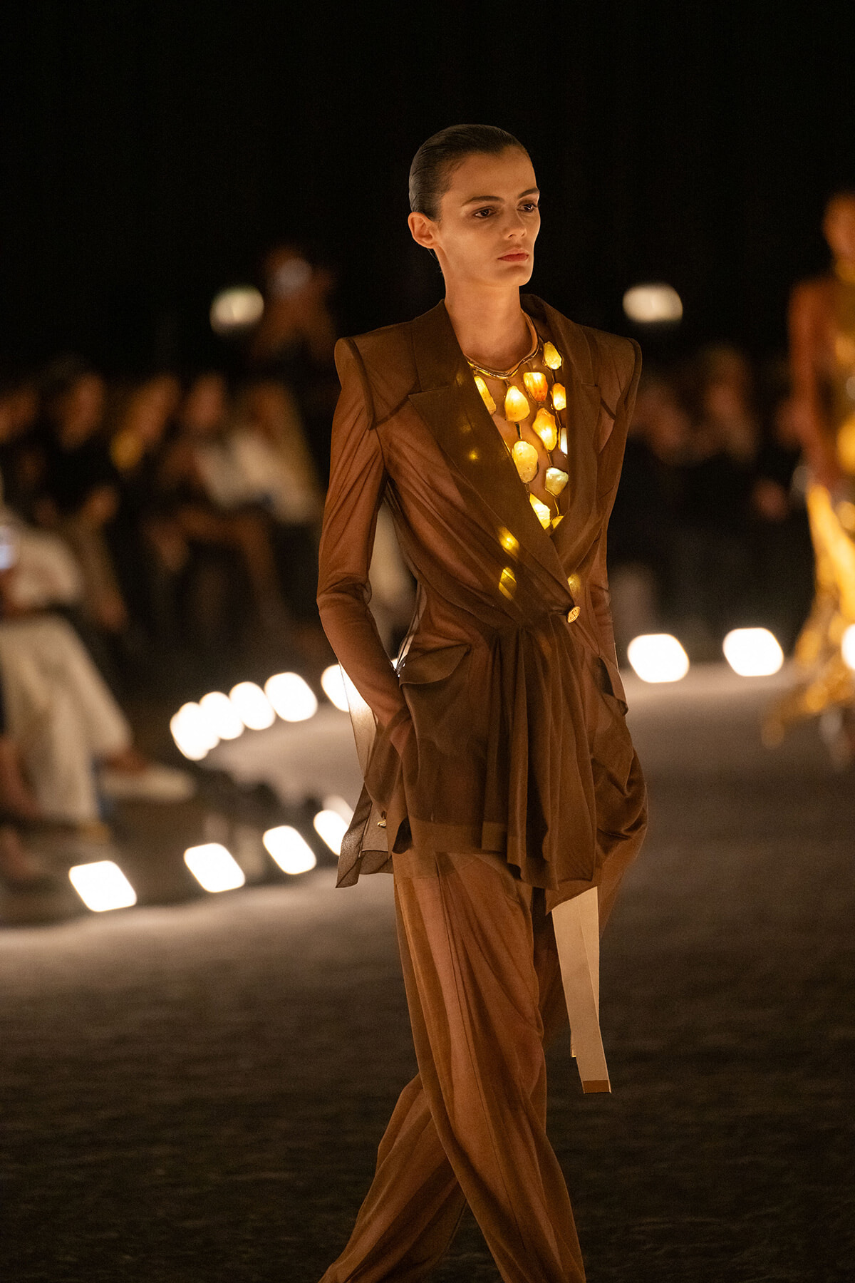 Model walks the runway at night in a brown suit with a layered gold statement necklace.