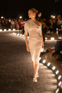 Model walks a dimly lit runway in a pale off-shoulder dress, audience blurred in background under warm lights.