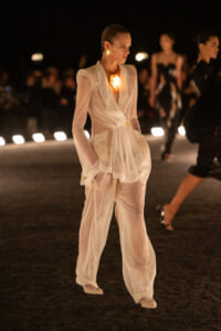 Model on a night runway wearing a sheer white blazer and wide-leg pants, with a large glowing pendant at the chest.