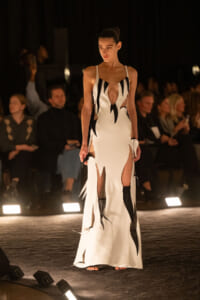 Runway model walks in a white gown with black flame-like cutouts and high slits, audience in the background under dim lighting.