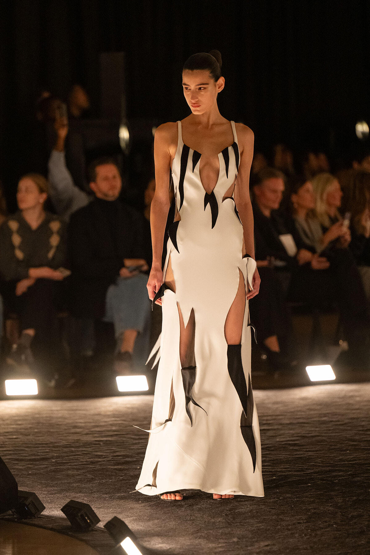 Runway model walks in a white gown with black flame-like cutouts and high slits, audience in the background under dim lighting.