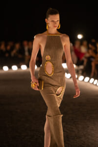 Model walks a runway in a taupe halter dress with large gold ring accents and matching jewelry in warm lighting.