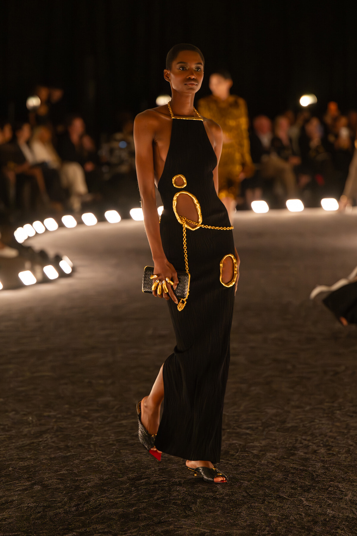 Model walks the runway in a black halter dress with oversized gold-ring cutouts and a gold chain belt.