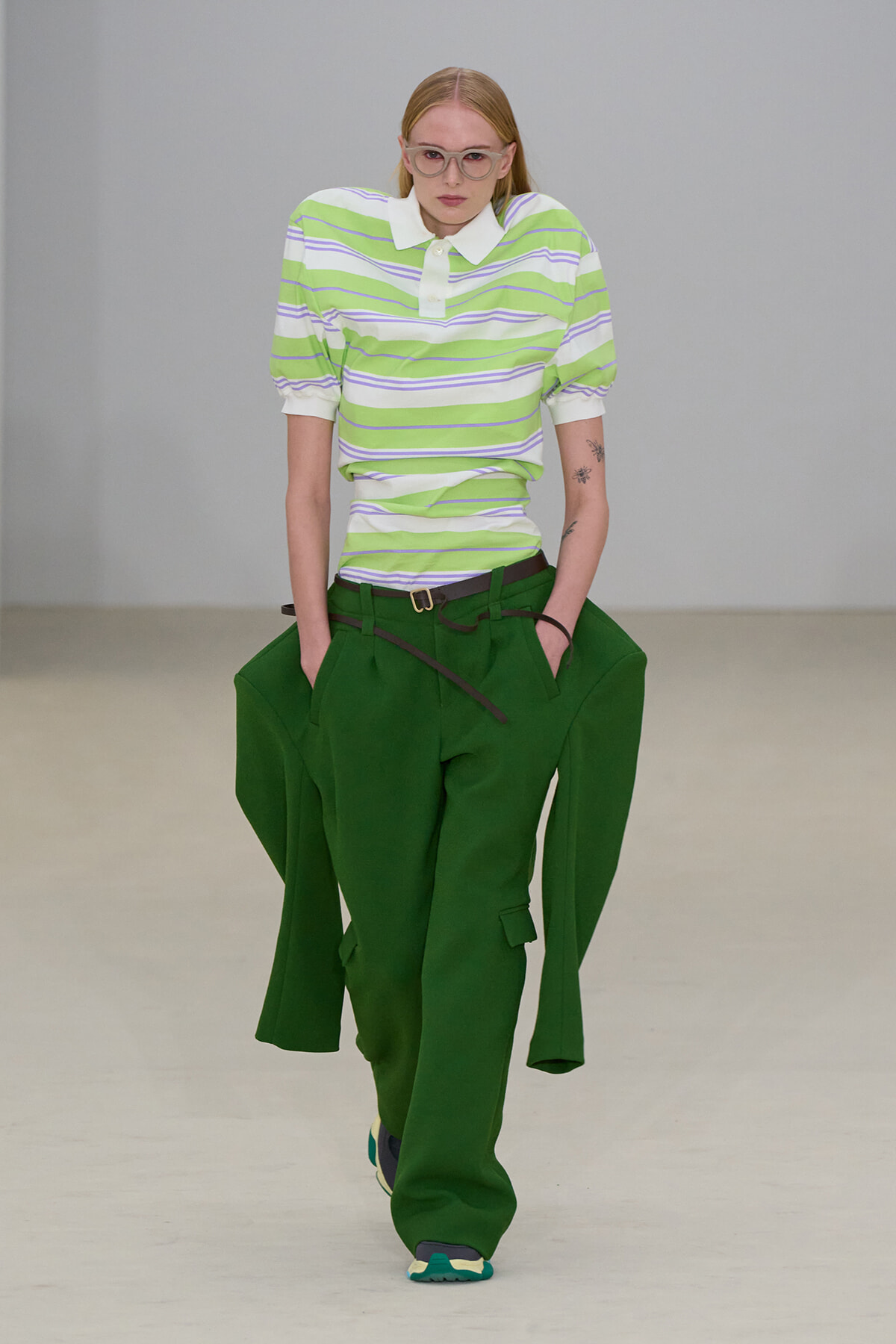 Model walking runway in lime-green striped polo and oversized green pants with a belt and sneakers, wearing glasses.