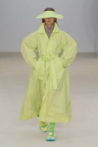 Model walks a run way in a neon lime trench coat with oversized sleeves and a matching belt, wide-brim hat completing the look; neon socks and colorful sneakers