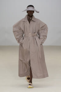 Model on a runway wearing a voluminous beige trench coat belted at the waist, with a wide-brim beige hat and pale sneakers.