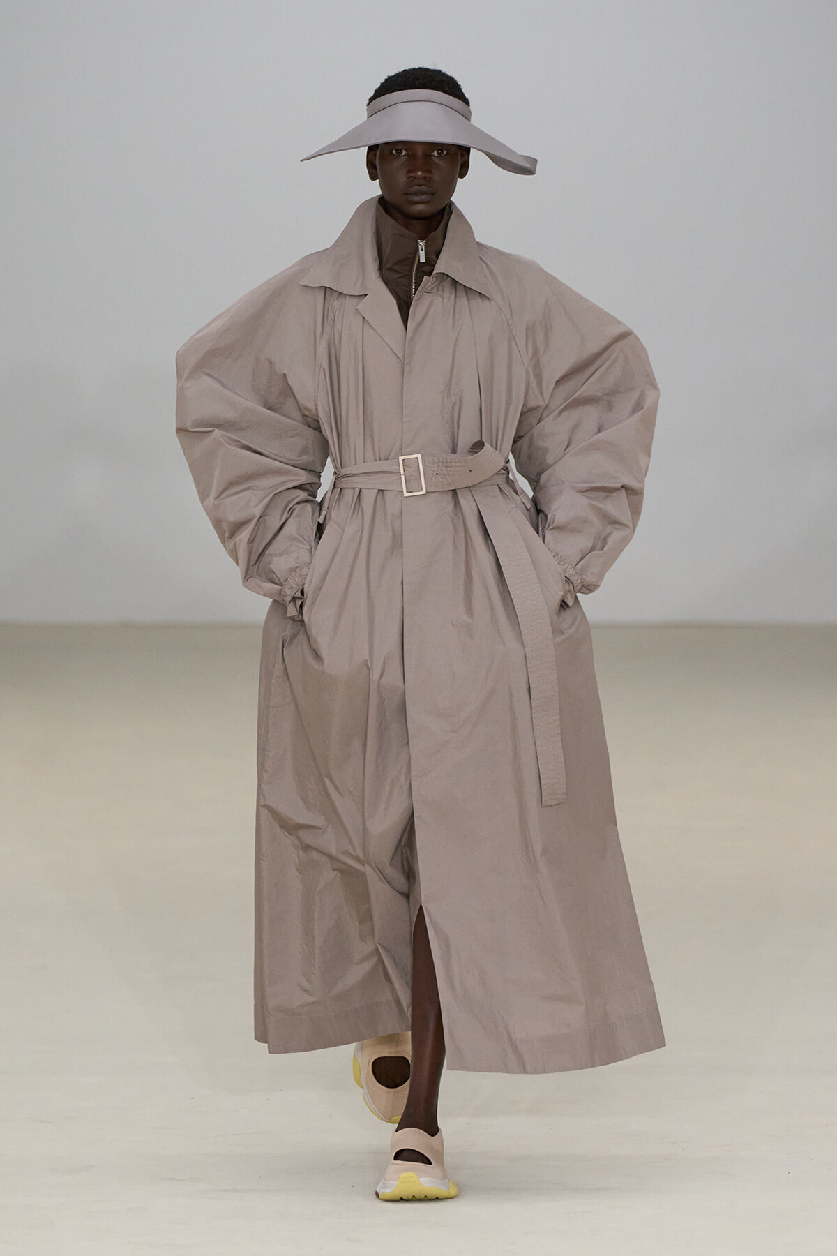 Model on a runway wearing a voluminous beige trench coat belted at the waist, with a wide-brim beige hat and pale sneakers.