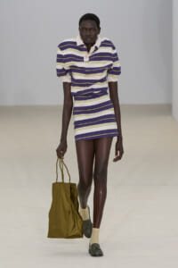 Model walking a runway in a purple, white, and yellow striped polo dress, carrying an olive tote bag at her side.