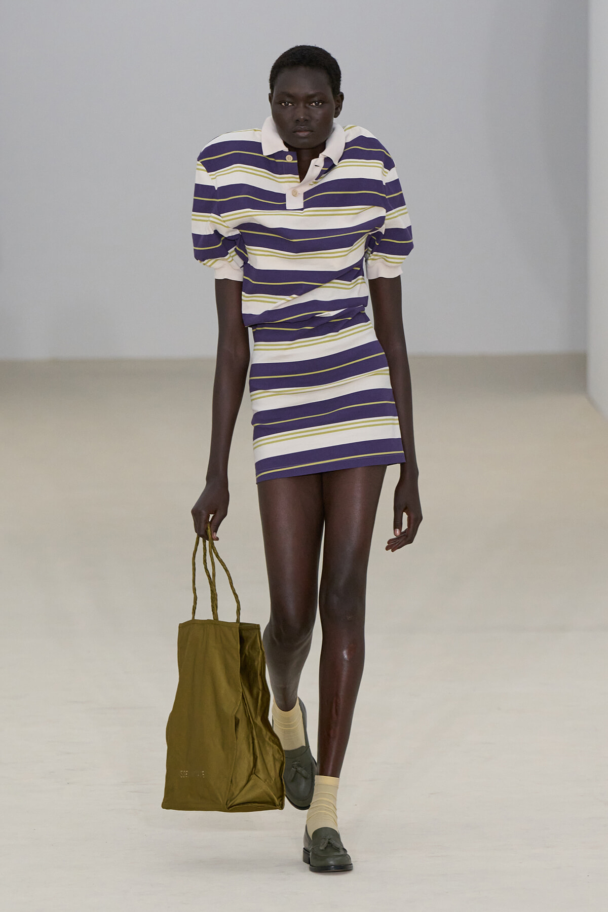 Model walking a runway in a purple, white, and yellow striped polo dress, carrying an olive tote bag at her side.