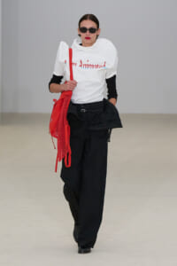 Female model in a white T-shirt with red lettering, black wide-leg pants, and a bright red mesh tote bag across the body, wearing sunglasses and red lipstick.