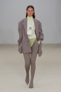 Model on a runway wearing an oversized lavender blazer over a pale lime turtleneck and lime bodysuit, with taupe ribbed tights and matching gloves; a white sculptural accessory is worn on the chest area.