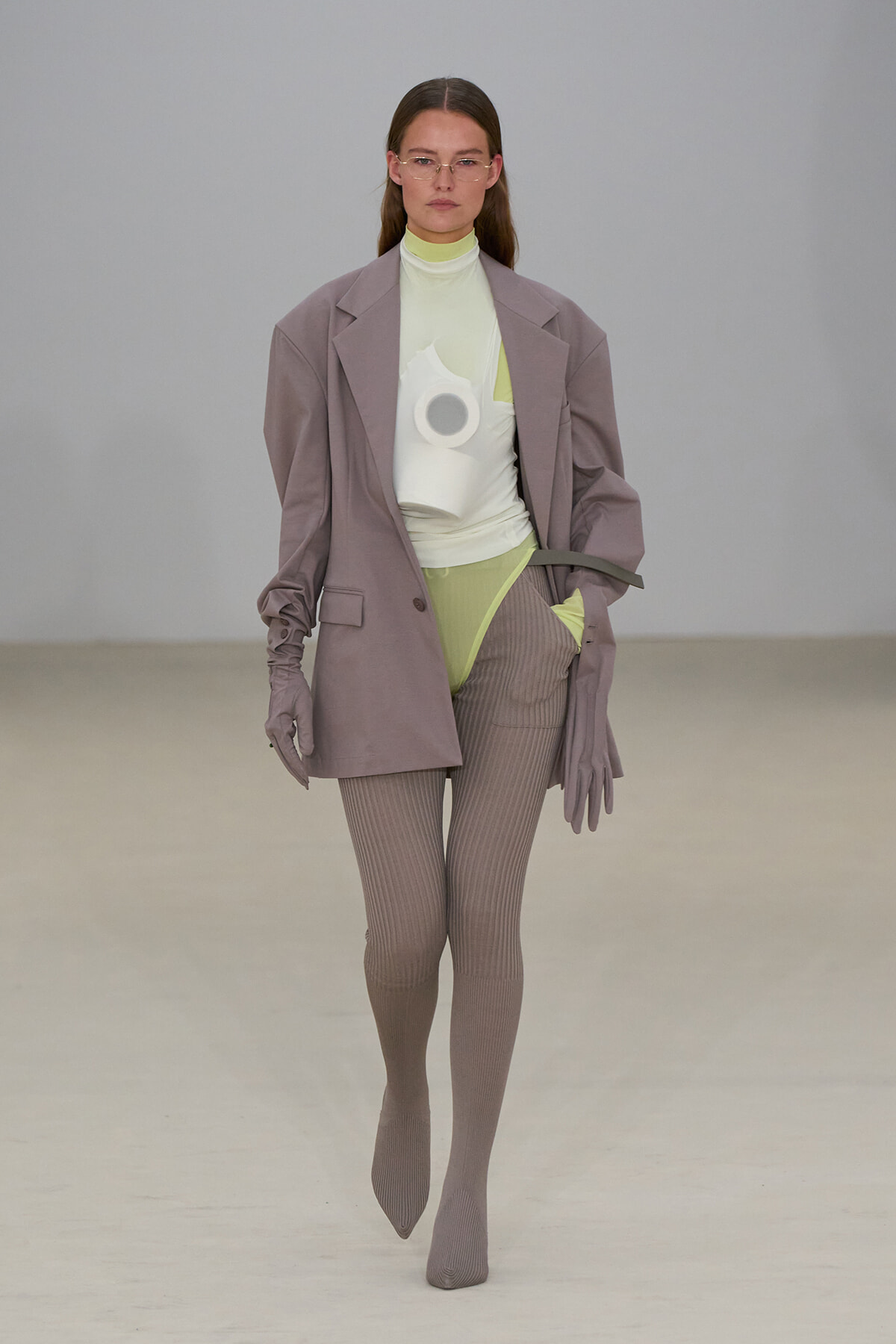 Model on a runway wearing an oversized lavender blazer over a pale lime turtleneck and lime bodysuit, with taupe ribbed tights and matching gloves; a white sculptural accessory is worn on the chest area.