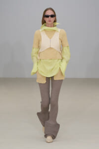 Model walking a runway in a pastel lime and beige layered ensemble with taupe ribbed leggings and oversized platform boots, plus dark sunglasses.
