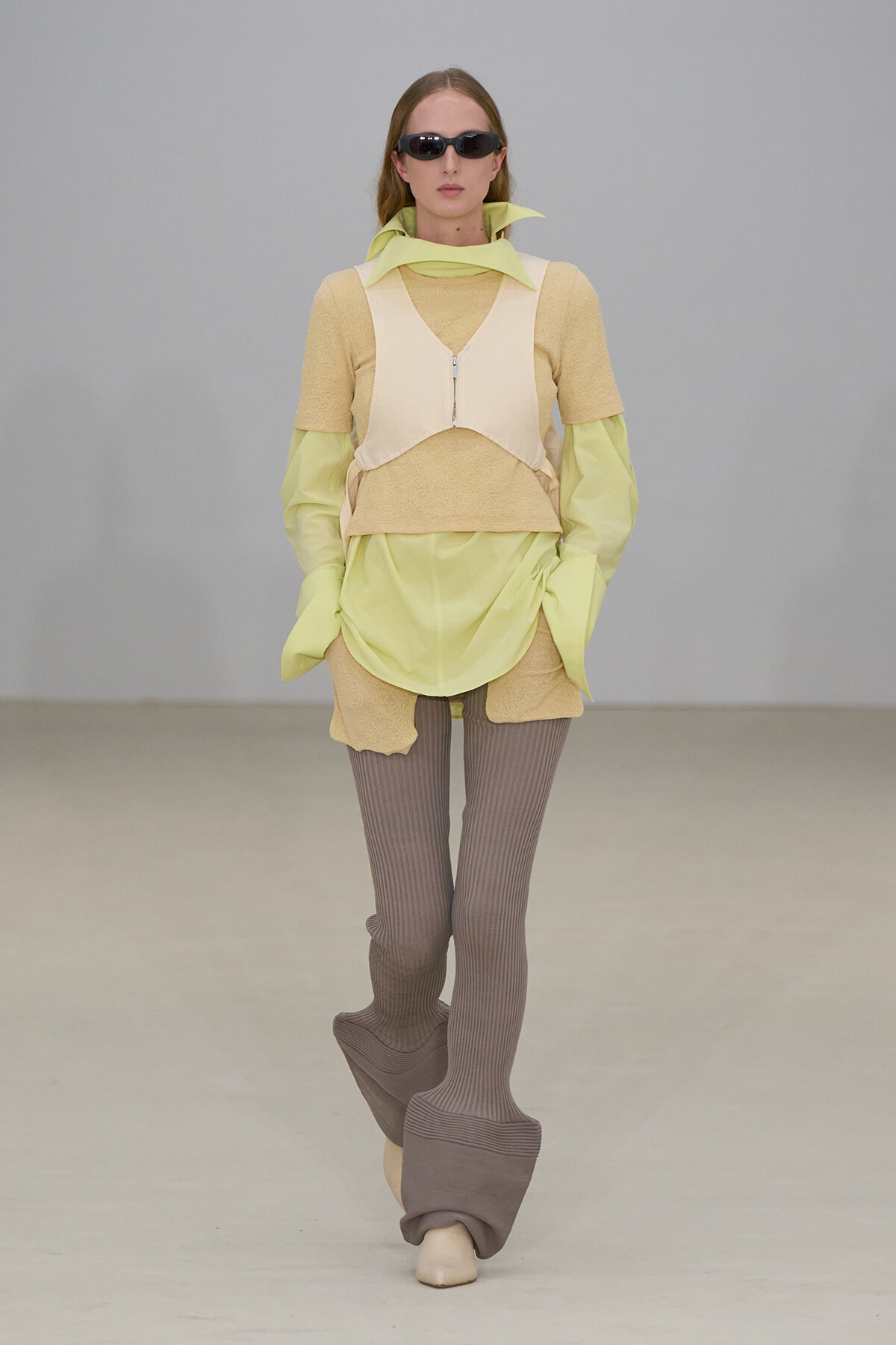 Model walking a runway in a pastel lime and beige layered ensemble with taupe ribbed leggings and oversized platform boots, plus dark sunglasses.