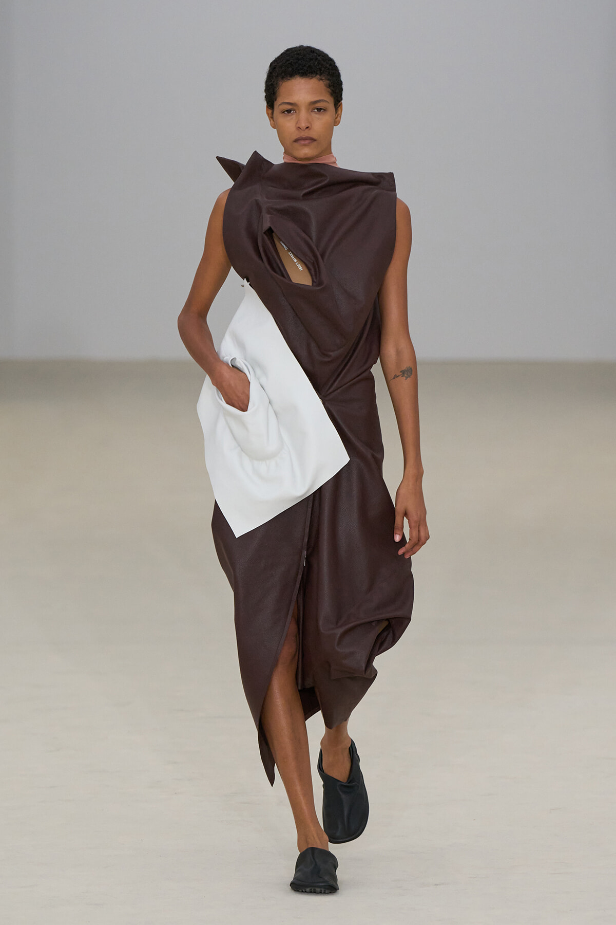 Model walks a runway in a dark brown asymmetrical dress with a large white pocket pouch at the waist; black slip-on shoes.