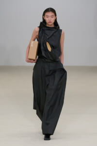 Model on the runway in a black sculptural sleeveless top with a circular chest cutout and wide-leg pants, carrying a brown cardboard box labeled CAMPER.