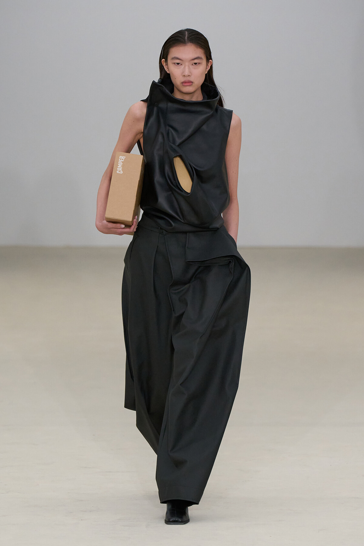 Model on the runway in a black sculptural sleeveless top with a circular chest cutout and wide-leg pants, carrying a brown cardboard box labeled CAMPER.