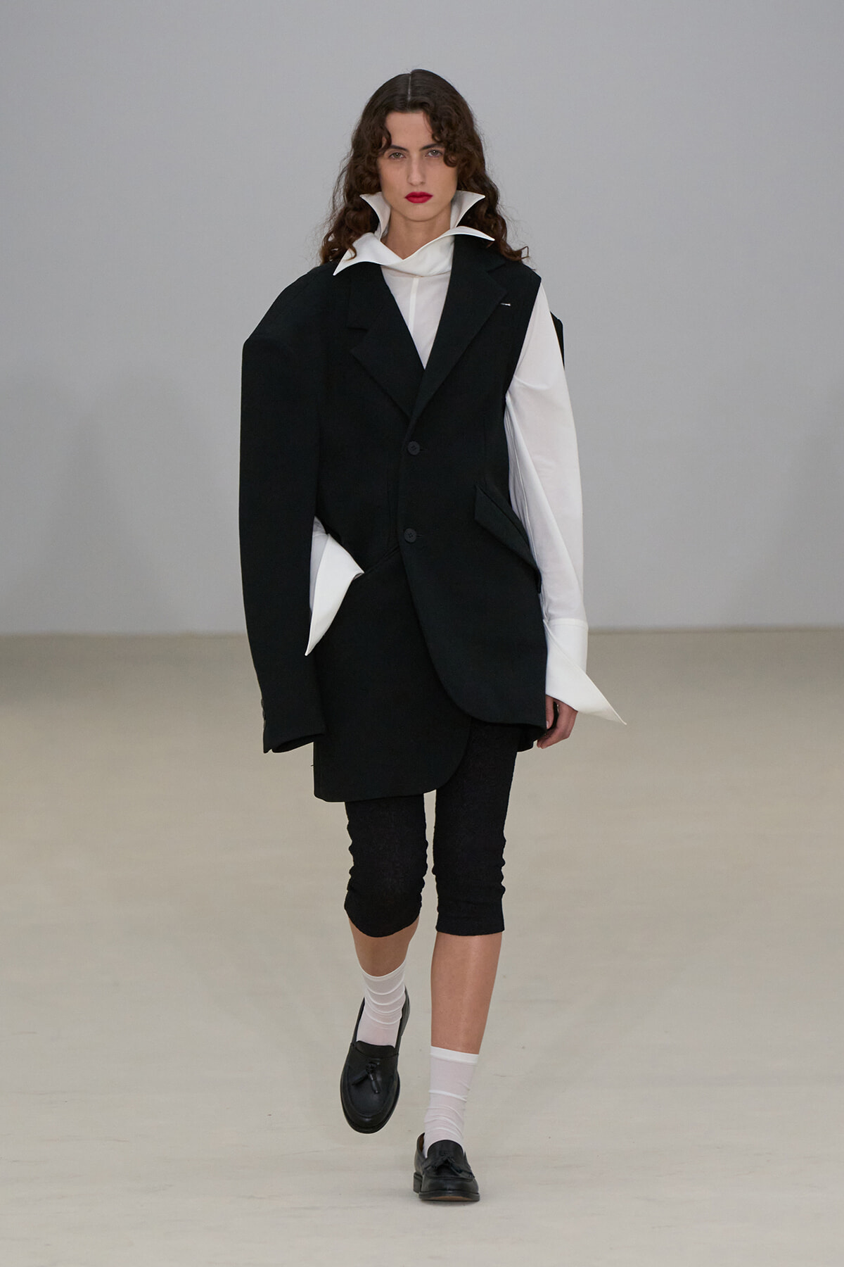Female model walking the runway in a black oversized blazer over a white shirt, with black cropped leggings and black loafers.