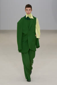 Model walking a runway in a bright green oversized suit with a pale lime shirt underneath, hands in pockets, minimalist backdrop.