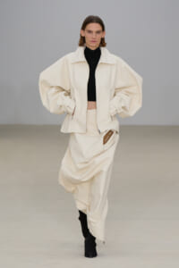 Runway look: oversized cream jacket with voluminous sleeves over a black cropped turtleneck and matching draped skirt, model walking forward.