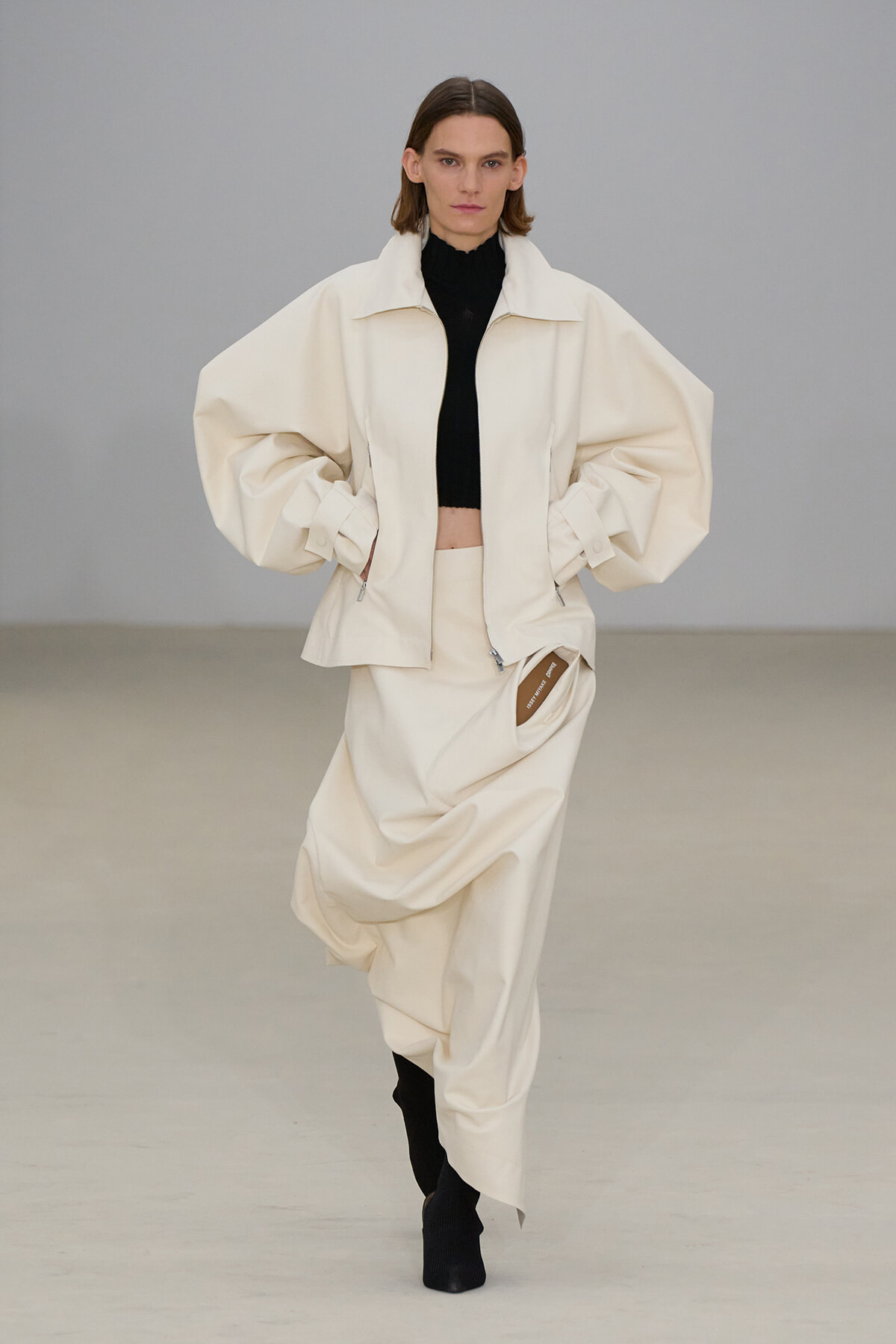Runway look: oversized cream jacket with voluminous sleeves over a black cropped turtleneck and matching draped skirt, model walking forward.