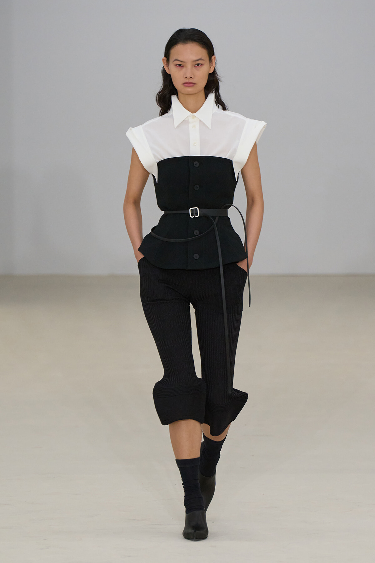 Model walking the runway in a white collared shirt paired with a black belted peplum vest and cropped black pants.